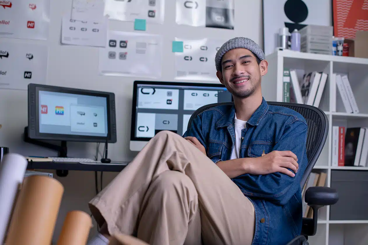 Male web designer smiles as he reclines into his chair, referring to designs on his Mac screens